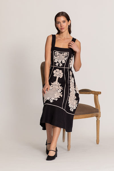 Woman wearing a black dress with white floral embroidery, standing against a plain background.